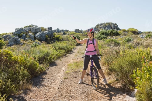 Preview: Female cyclist straddling purple mountain bike on trail with helmet and backpack holding smartphone