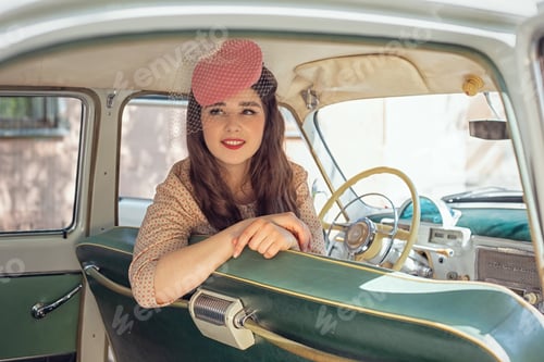 Preview: A beautiful happy girl in a small retro hat and dress sits in a retro car.