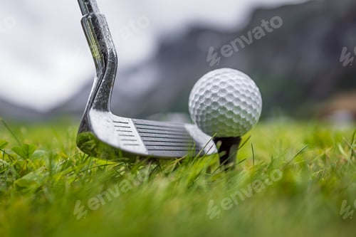Preview: Golf club and ball on wet green field with mountains in background.