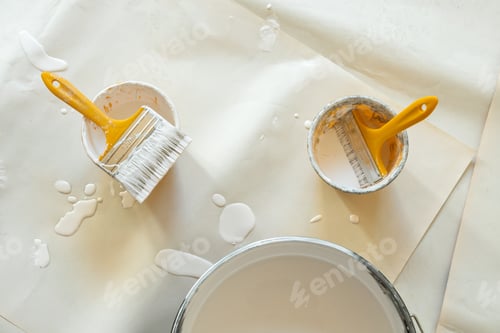 Preview: Two paintbrushes with yellow handles rest in small plastic cups filled with white paint