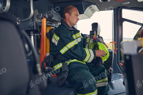 Preview: Photo of adult fireman with helmet in hands sitting in fire engine