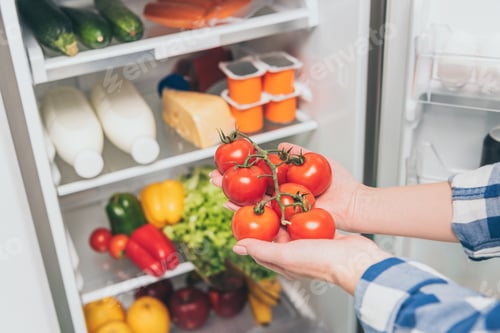 Preview: cropped view of woman holding tomatoes near open fridge with fresh food on shelves