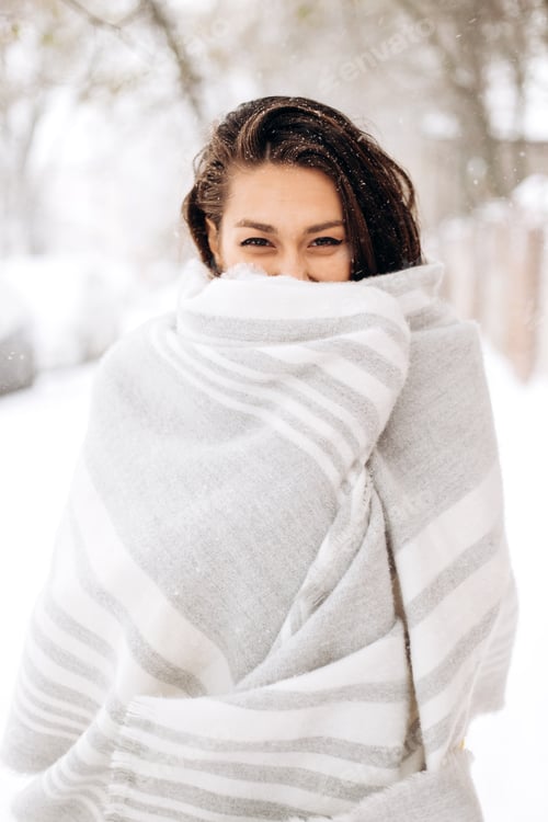 Preview: Charming dark-haired girl wrapped in a gray scarf is standing in a snowy street on a winter day