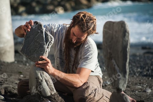 Preview: Young hippie with dreadlocks makes weightless balancing sculptures from stone on the seashore
