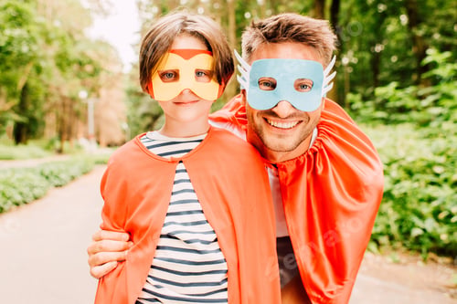 Preview: happy father and son standing in red cloaks and superhero masks, smiling and looking at camera