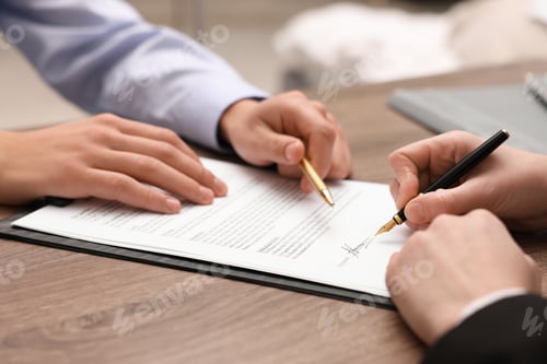 Preview: Man pointing at document and woman putting signature at wooden table, closeup