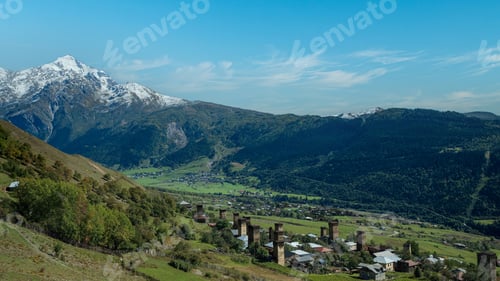 Preview: Historic mountain village with medieval towers beneath snowy peaks and bright blue sky.