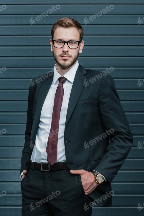 Preview: Portrait of handsome young businessman in eyeglasses standing with hands in pockets and looking at