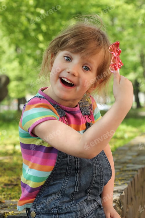 Preview: Cute little happy smiling girl holding and showing her red lollipop with green summer background