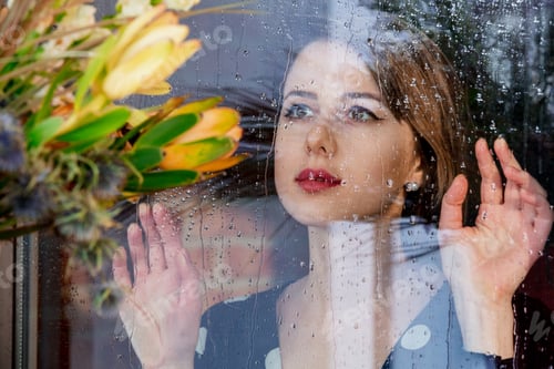 Preview: woman at the wet window after the rain misses the plants standing outside the window.