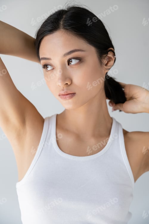 Preview: pretty asian woman in white tank top touching brunette hair and looking away isolated on grey