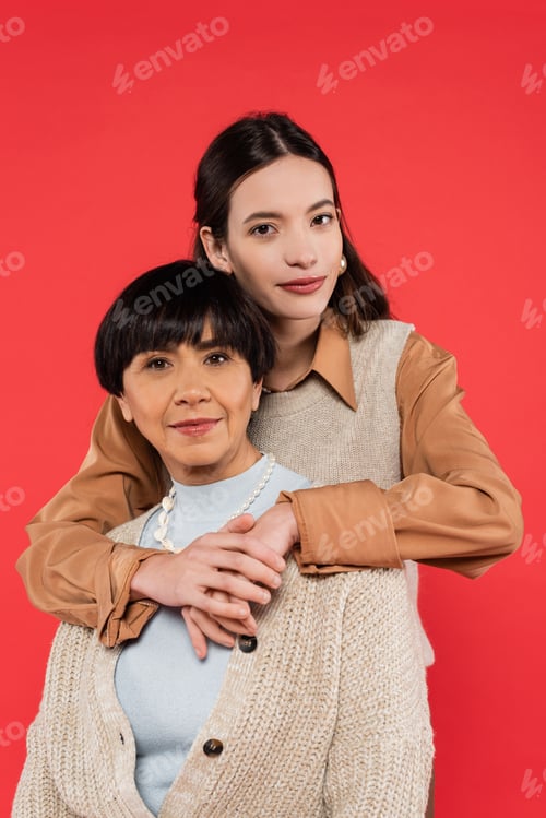Preview: young asian daughter in trendy clothes hugging mother isolated on coral
