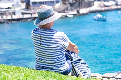 Preview: Rear view of senior caucasian woman in blue hat sitting at the harbour admiring the sea