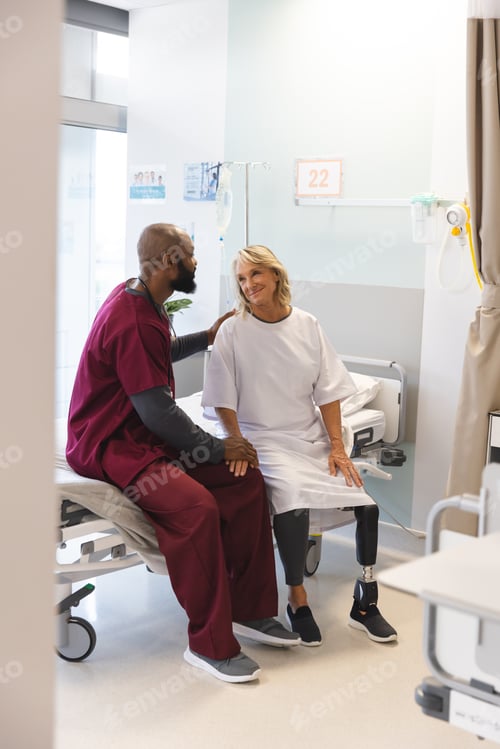 Preview: Senior caucasian female patient with prosthetic leg and african american male doctor sitting on bed
