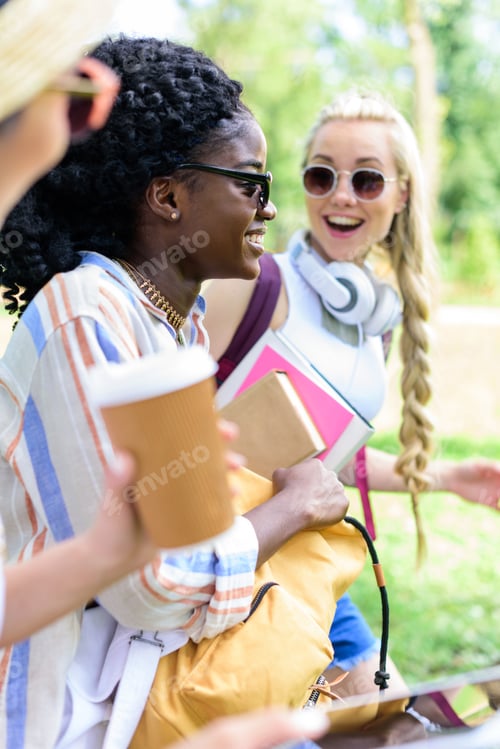 Preview: smiling young multiethnic students holding books and paper cup while talking in park