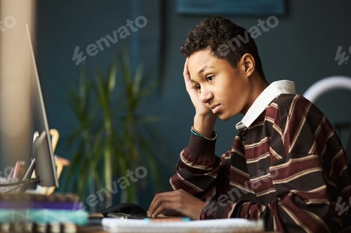 Preview: Teenage Black Boy Sitting at Desk Using Computer and Looking Thoughtful