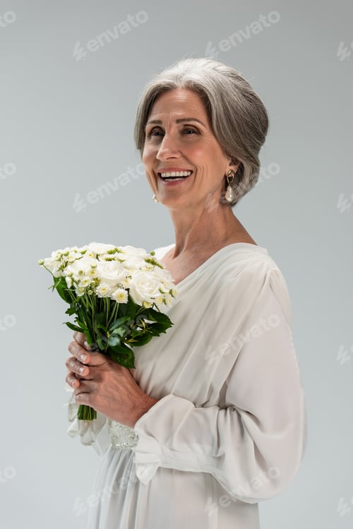 Preview: joyful middle aged bride in white wedding dress holding bouquet isolated on grey