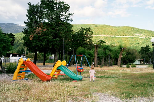 Preview: Kid walks along the playground, past the slides to the swing. Back view