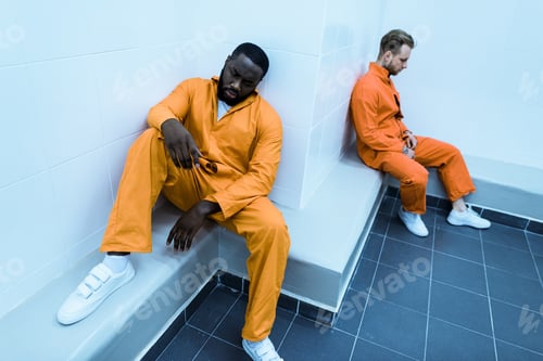 Preview: high angle view of multicultural prisoners sitting on benches in prison cell