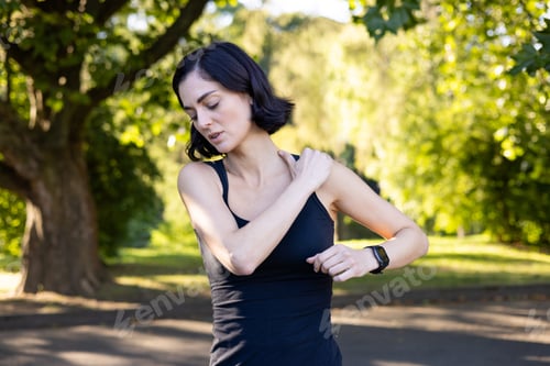 Preview: A close-up photo of a young woman doing sports in the park, standing and holding her hand by her
