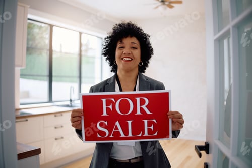 Preview: Happy real estate agent holding sing 'for sale' and looking at camera.