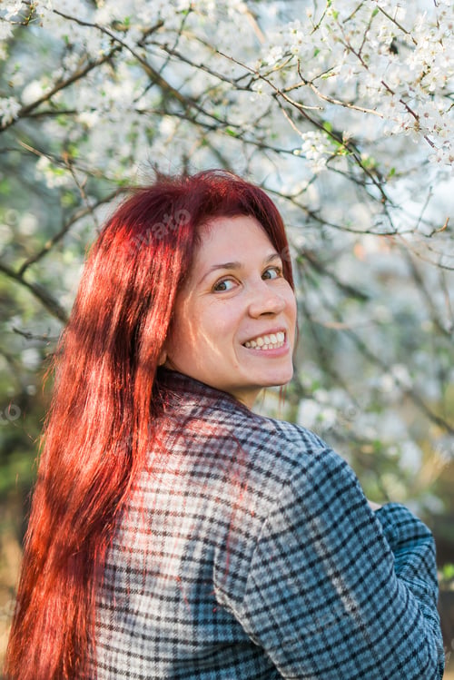 Preview: Beautiful red-haired woman enjoying smell in a flowering blooming spring garden. Spring blossom