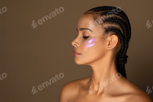 Preview: Sensual black middle aged woman with perfect even healthy skin using eye patches, brown background