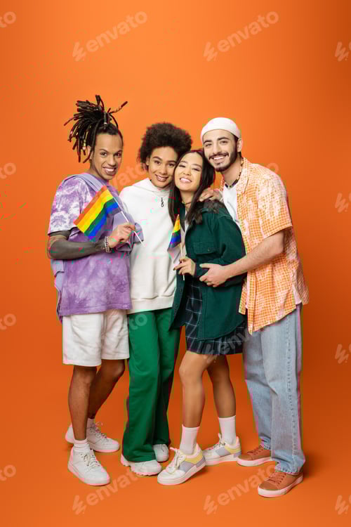 Preview: full length of multiracial friends in stylish outfit holding lgbt flags on orange background