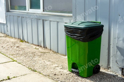 Vista previa: Contenedor de basura de plástico verde parado en la calle