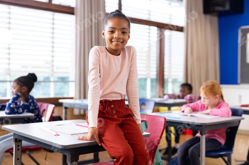 Preview: Smiling girl sitting on desk in classroom, students writing in background at school