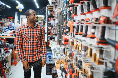 Preview: African american man holding and choosing electric screwdriver, standing in construction store