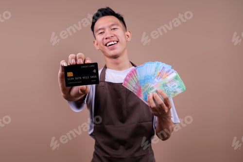 Preview: Attractive Young Asian Man In Brown Apron Showing Credit Card And Holding Indonesian Rupiah Bills