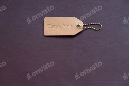 Preview: Flat lay of a light brown leather tag with Thank You word on dark brown background