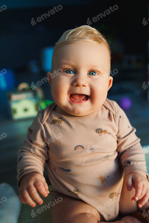 Preview: Fair-haired boy with blue eyes baby 11 months in a bodysuit sits on the floor at home and smiles