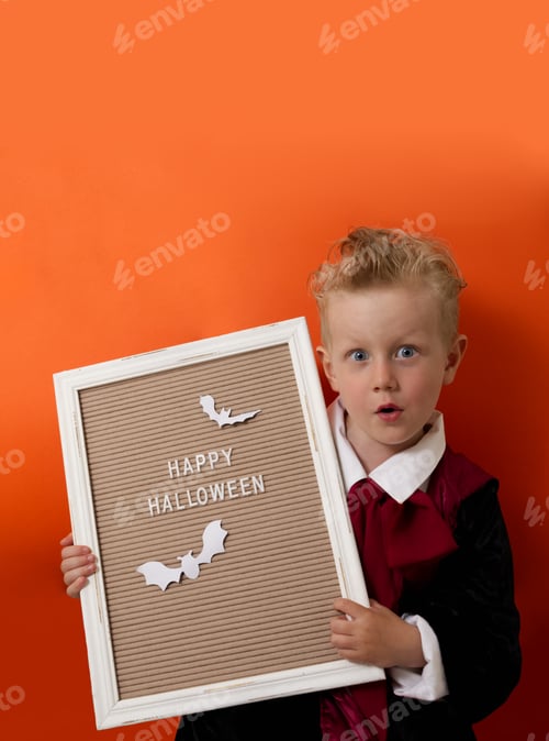 Preview: A boy in a vampire costume holds a board with a wish for a happy halloween on an orange background
