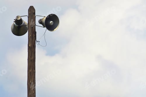 Preview: Public address horn loud speakers on wooden pole over blue sky.