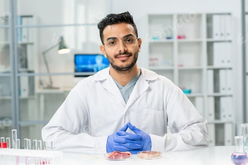 Preview: Young successful scientific researcher in whitecoat looking at you by workplace