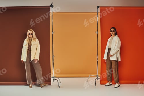 Preview: autumn fashion, stylish multiethnic models in sunglasses and outerwear posing on colorful backdrop