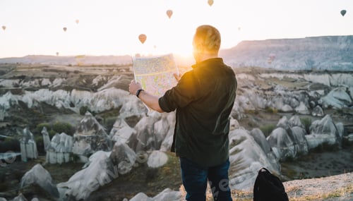 Preview: Unrecognizable traveler looking at map with air balloons over highlands in background