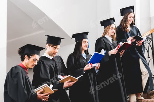 Preview: happy students in graduation gowns holding diplomas and reading books