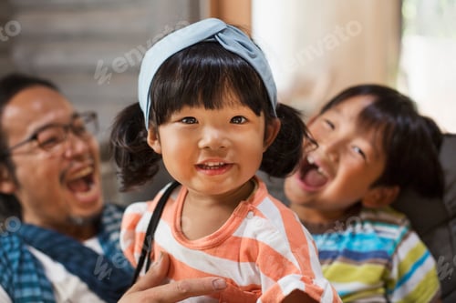 Preview: Man, boy and young girl with black pigtails wearing blue hairband sitting on a grey sofa, laughing.