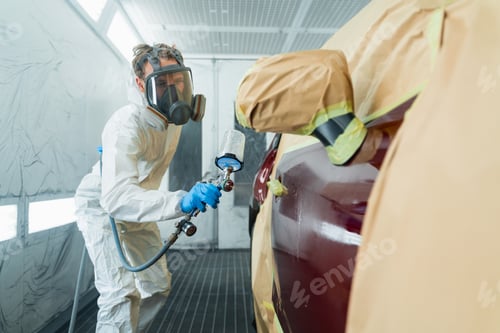 Preview: Auto painter in a protective suit and mask sprays thinner or other cleaner on car door