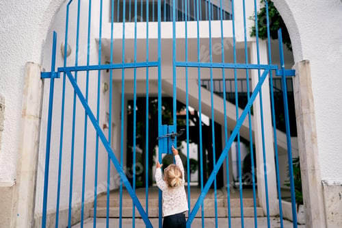 Preview: Little girl is trying to open the closed metal gate of the house while standing in the yard. Back