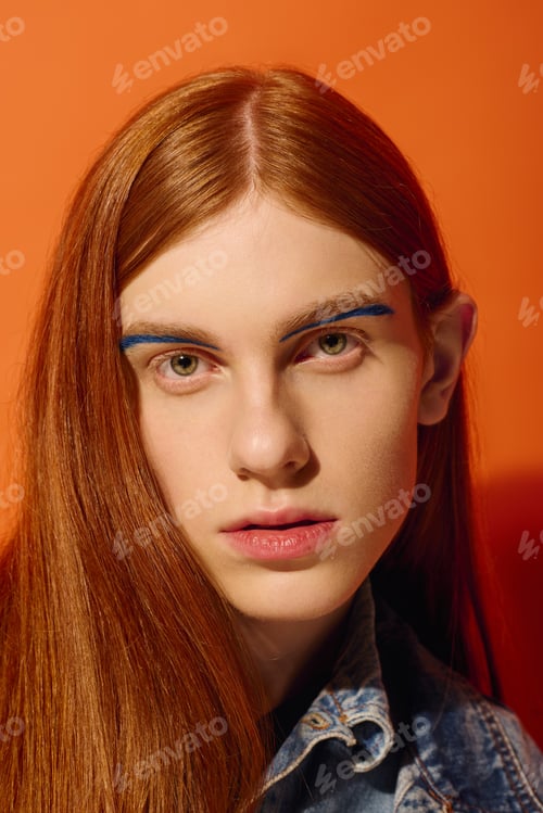 Preview: Emotional young man with long red hair and striking blue eyebrows against a vibrant orange backdrop