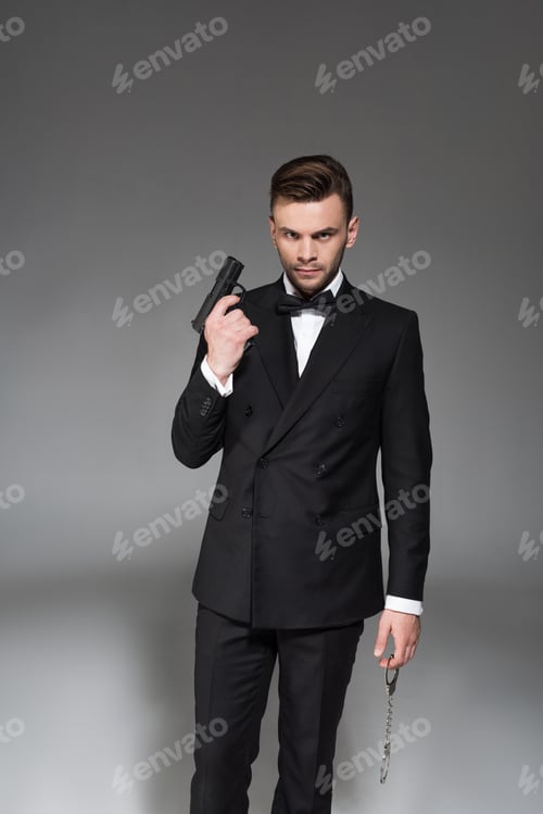 Preview: handsome secret agent in tuxedo holding handgun and handcuffs, isolated on grey