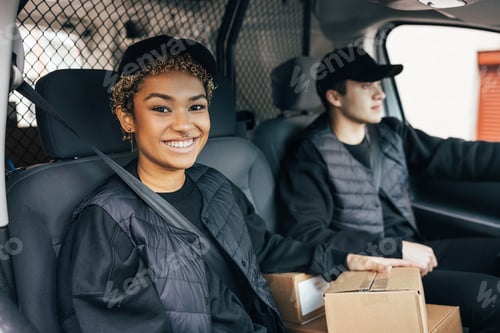 Preview: Smiling woman in uniform sitting in delivery van and looking at camera while her coworker driving