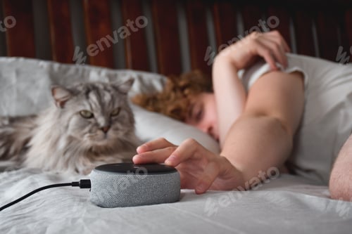 Preview: Young man sleeping in bed with cat and pressing button on virtual assistant to stop alarm