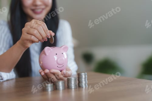 Preview: Smiling businesswoman saving money putting coins into piggy bank sitting at desk