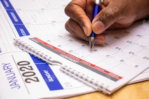 Preview: african American man using blue pen to write on a January calendar doing events and planning