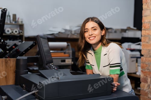 Preview: cheerful woman smiling near printer machine while working in print center